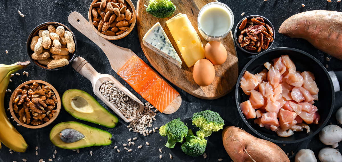 Biotinhaltige Lebensmittel wie Lachs, Geflügel, Avocado, Milchprodukte, Brokkoli, Bananen und Nüsse in Schüsseln und auf einer Schieferplatte.