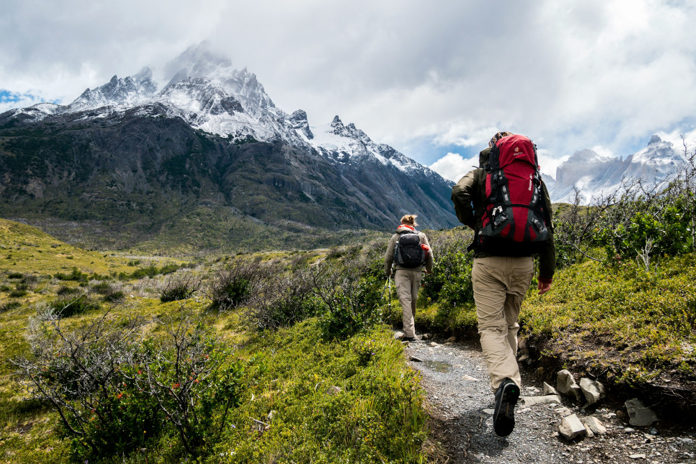 Zwei Wandernde auf Steinpfad zwischen Grün, unterwegs Richtung Gebirge mit weißer Spitze, raue Umgebung