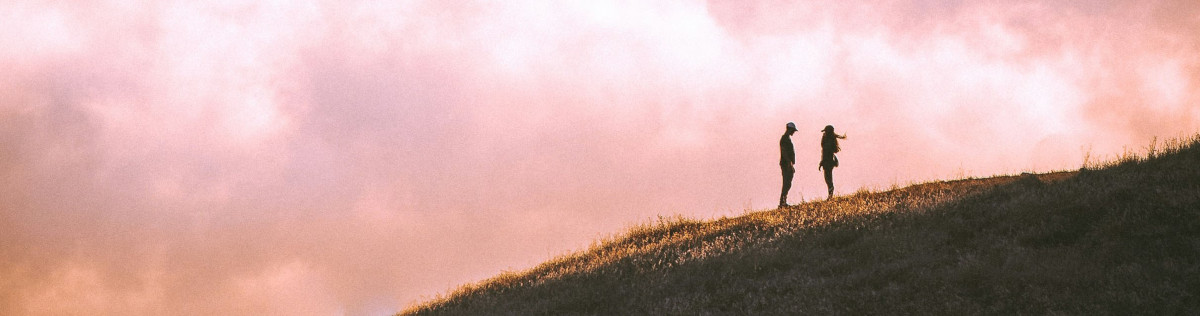 Foto: Ein Paar als schemenhafte Gestalten auf einem Hügel, Abstand zwischen ihnen, der Himmel im Hintergrund blassrosa