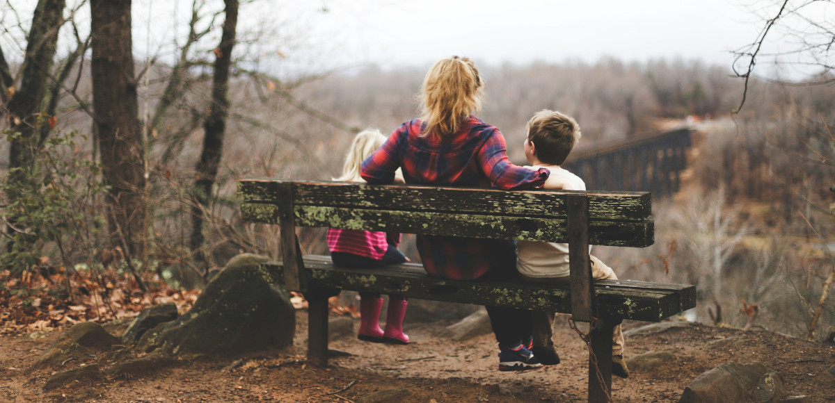 Foto: Blick von hinten auf einzelnes Elternteil mit zwei Kindern auf einer Bank, im trostlosen Herbstwald