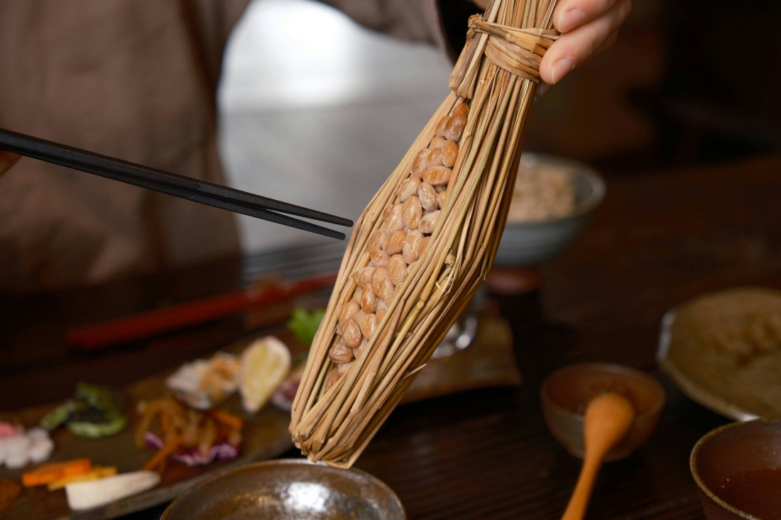 Natto im Strohmantel wird über eine Schüssel gehalten. Zwei Stäbchen sind in der Bewegung, um das Natto in die Schüssel zu schieben. Im Hintergrund verschiedene Gemüssesorten, in kleine Stücke geschnitten