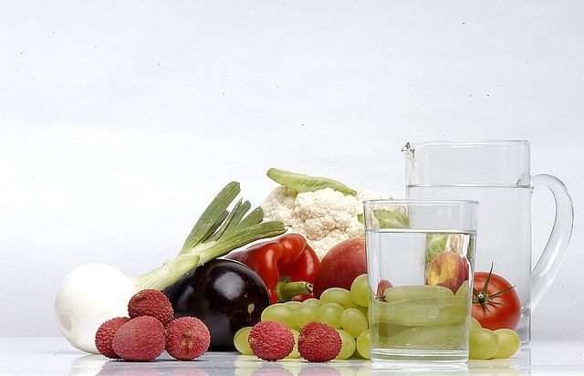 Foto von Obst und Gemüse wie Litschis, Trauben, Tomaten, Paprika, Fenchel, Aubergine und Blumenkohl neben einer Karaffe und einem gefüllten Wasserglas