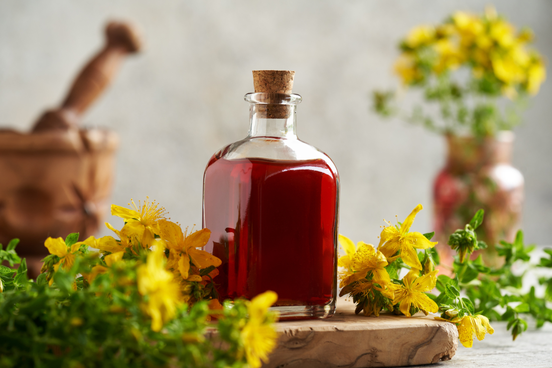 Rotöl in einer Flasche auf einem Holzbrett, umgeben von Johanniskrautblüten.